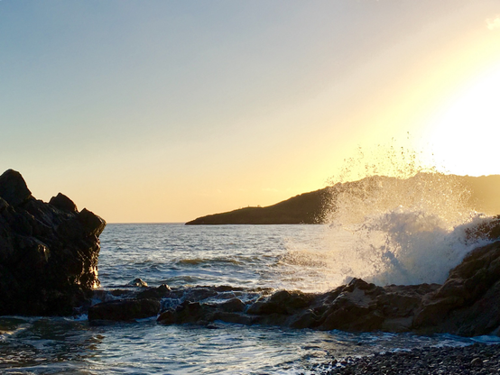Blue and apricot sky over Langland Bay with a wave crashing high against rocks in the foreground. Pic taken from Rotherslade Bay at high tide.