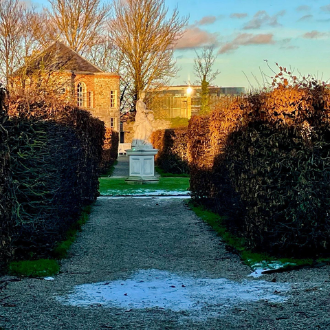 Formal garden path. Sculpted box hedges to side. Patches of ice on ground and lawn. A classical statue in mid ground. A French-style chateau and modern glass building in background lit by golden setting sunset.