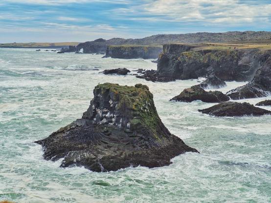 Lava rock formations with moss growing on top in the sea near a coast with rocky cliffs. The wild green sea is foamy from the waves.