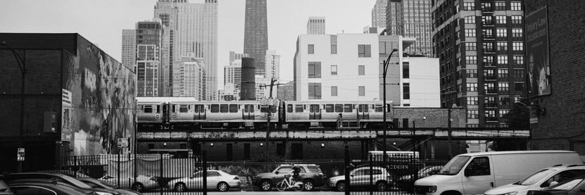 A cyclist rides a bike down a street lined with cars while a train crosses above on an elevated train track. Buildings tower over the elevated train.