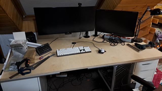 Eine helle, unbehandelte Holztischplatte mit zwei Monitorhalterungen, an denen jeweils ein Monitor befestigt ist. Darauf eine Tastatur, Mouse, Lautsprecher, etwas Werkzeug und Akkuschrauber.
Vorne unter der Tischkante sind dünne Aluminiumprofile montiert.
Links und rechts liegt die Tischplatte auf weißen Schränken auf.