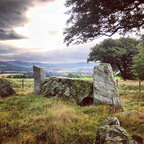 The very large recumbent stone and two flankers of an RSC, with a view acorss the valley to distant hills. There are grey clouds with the sun breaking through.