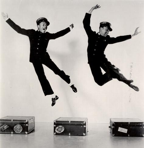 The brothers leaping high in a dance pose above a row of suitcases
