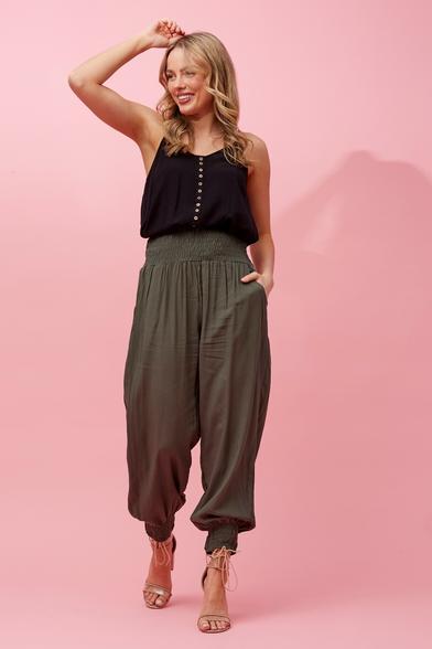 A blonde white woman wears a black sleeveless round-necked top with contrasting tiny ivory-look buttons, tucked into a pair of drapey khaki pants with a wide shirred elastic waistband and shirred elastic ankle cuffs. Her left hand is tucked into the pants' side pocket. She wears ugly beige high-heeled sandals with elaborate fussy string ties around the ankles, which are an awkward mismatch with an ankle-focused pair of pants