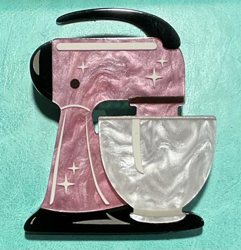 An acrylic brooch of a vintage stand mixer.
This acrylic brooch of a vintage stand mixer has a black handle, base, and rear with grey highlights. The body of the mixer is pink swirled acrylic with white star highlights and white stripes. The bowl of the mixer is white pearl acrylic with white striped highlights. The brooch is resting on a teal green background.