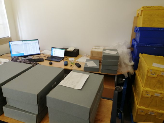 A messy office environment with grey and brown storage boxes for medieval and early modern charter index slips, a laptop with attached screen, and blue and yellow moving crates to the right side.