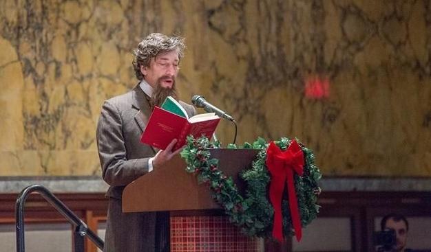 Image shows Neil Gaiman in a fake beard, stood in front of a lecture – which is adorned in a festive garland and red ribbon. Neil is reading Charles Dickens' A Christmas Carol in the New York Public Library