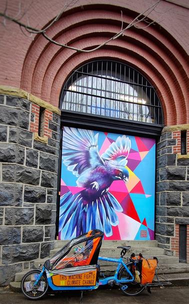 Electric cargo bike with kid in front box. Beautiful mural with a bluebird with multicolored triangles and background. Ebike is also blue and called the bluebird.