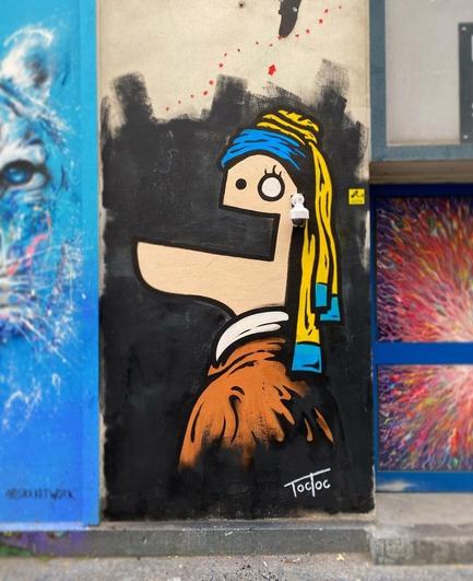 Street art wall: cartoon style. A girl, painted on the side, looks in the direction of the viewer, laughing with an oversized mouth. She is a cartoon version of the painting "The Girl with the Pearl Earrings" by Vermeer. Instead of earrings, she wears a security camera.