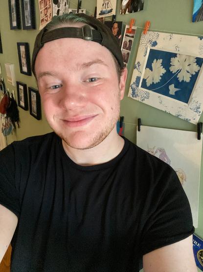 A selfie of Haven, a white man wearing a black t-shirt and a backward gray hat. He has teal hair. Behind him is a wall of small art pieces hanging from twine.