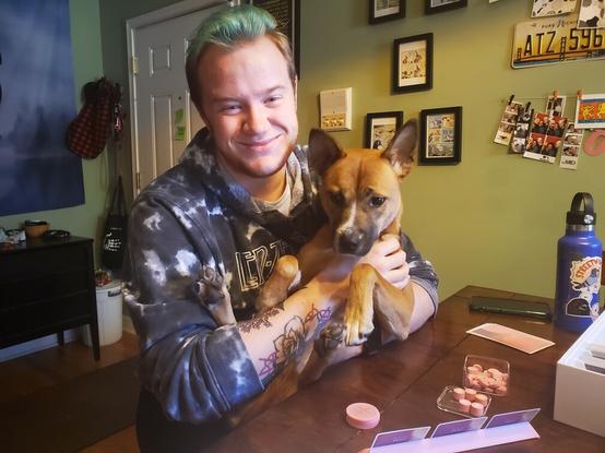 A photo of Haven holding his dog, Erin, who is a medium-sized tan-colored dog who is half New Guinea Singing Dog. He is sitting at a table playing the board game Fog of Love.