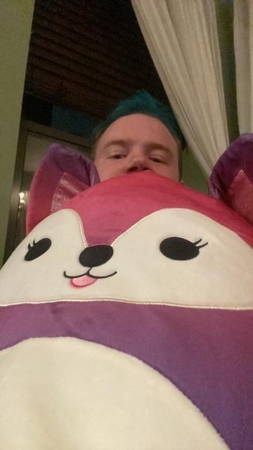 A selfie of Haven holding a large pink, purple, and blue fox Squishmallow that takes up most of the frame. His eyes are peaking over the top of the Squishmallow to look at the camera.