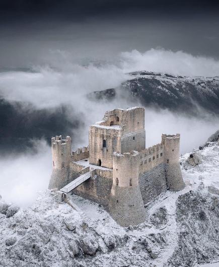 This image is by Franco Nicolli (IG: franconic89).

Atop a snow-draped mountain ridge stands a fortress ruin. Its high walls form a square bastion around a central tower. Each corner is marked by a circular, crenelated tower.