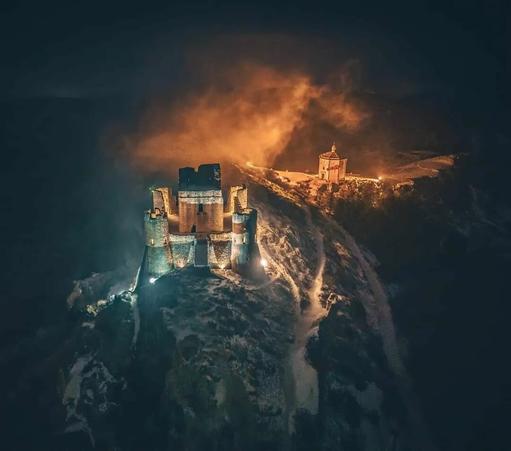 This image is by Mauro Pagliai (IG: Mauro_Pagliai):


Night has fallen on the ruined keep. The lights that illuminate its walls and nearby chapel bleed into the night fog, creating a strange diffuse effect like fire and smoke.