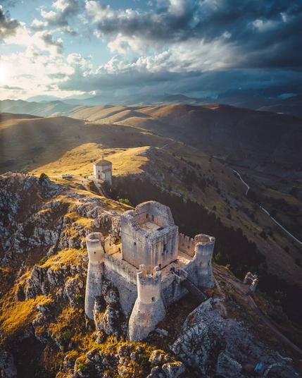 The photo is by Mauro Pagliai (IG: Mauro_Pagliai).

As the sun shines above the horizon, the blue sky falls to waves of marching clouds. Despite the growing dark above, the rocky peaks and grassy hills are touched by golden sunlight.

In the fore of the frame stands the keep, its base incorporating the rocky slopes and ridges into its construction.