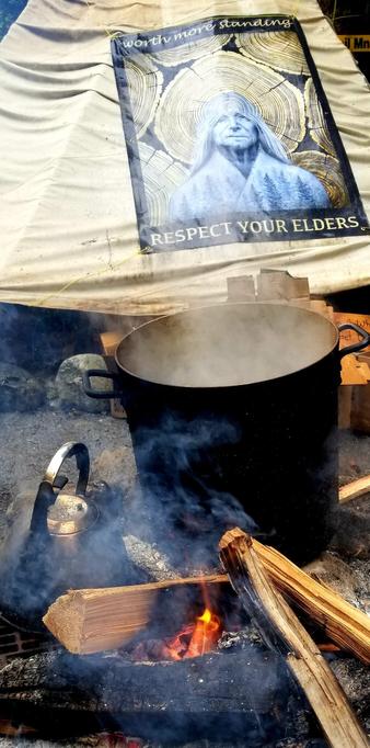 A big pot of boiling water, atop a campfire with a tipi in background, with sign saying: Respect Your Elders