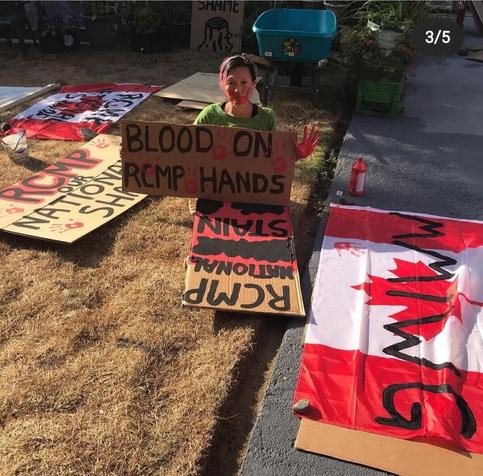 Me holding up handmade sign saying: Blood on RCMP hands. Red handprint across my face, holding my left hand up, covered in red paint.  An array of anti-RCMP protest signs surround me.