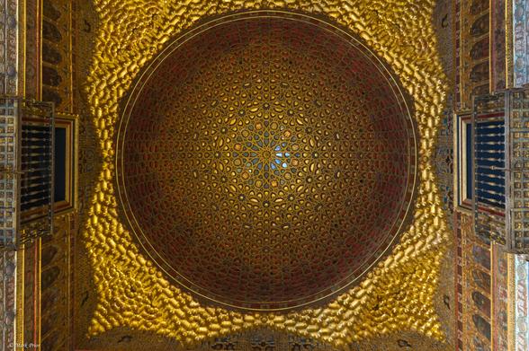 Standing in a square room looking up at a golden circular vaulted roof with an intricate pattern. The central element of the half dome is a 12 pointed star. Within the lattice work around the central star are images of rampant lions and castles. A gilded roof surrounds the dome and extends to the walls of the room. On the walls on either side are two false balconies.