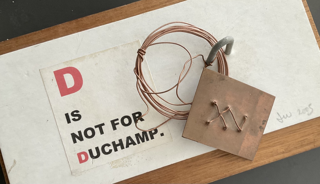 A statement: “D is not for Duchamp”. Written on white card with a copper wire coil Criss cross woven into a flat rectangular copper plate fixed to a wooden base.