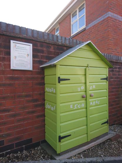 A small shed/cupboard, wider than it is deep and the height of the red-brick garden wall it backs onto, with full-width doors opening forwards and a gable roof. It is built in shiplap with black metal hinges and covered in roof felt. It is painted lime-green, with the words BOOKS and BOOK EXCHANGE stencilled in white on the side and front. The surrounding ground is covered in gravel, and there is a red-brick house behind the wall.