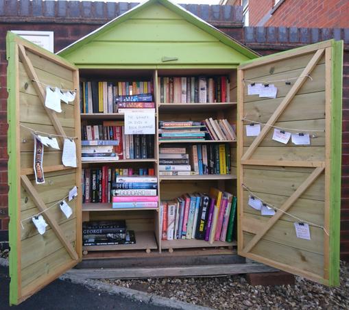The book exchange with its doors open wide, showing shelves of books. On the inside of the doors there are bulldog clips hanging on string, and many hold jokes written on small pieces of paper.