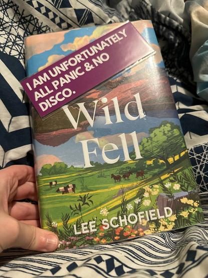 Hardcover copy of Wild Fell by Lee Schofield, with a purple bookmark on top of it which reads “I am unfortunately all panic & no disco”.