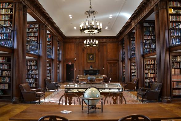 A large reading room with deep, luxurious wooden columns and paneling. Circular chandeliers, decorated in zodiac symbols hang from a high ceiling. Most notable is that two floors of shelving are exposed on either side of the room, accessible from stairs not visible in the photo. The second level overlooks the main room by balconies fenced with wrought iron. Long tables and leather chairs are in the middle of the room. A portrait is centered at the far wall over a bust of Dante. The room looks quiet, and comfortable, but still formal. You could lose yourself for a few pleasant hours perusing books on the shelf or delving deeply into your research.