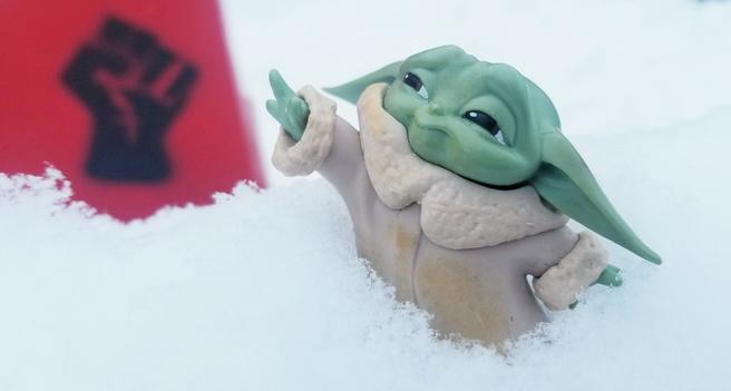 A spray painted solidarity fist in black on red newspaper box on the left.  Baby Yoda reaching up towards the solidarity fist, on the left. Surrounded by snow.