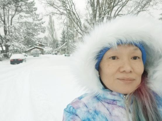 Selfie with my furry Winter parka hood up, snowy road in background with trees & houses.