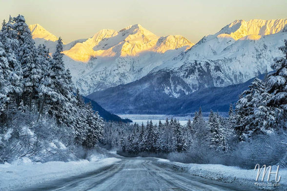A shot of the scenic Seward highway in Alaska shows snow-capped mountains in the background and snow-draped trees in the foreground.