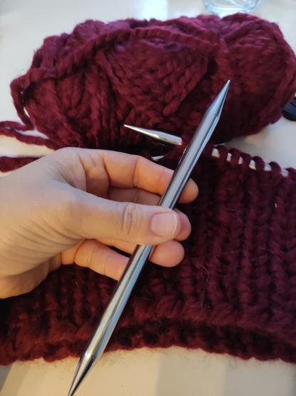 Hand hält eine sehr dicke Stricknadel. Im Hintergrund ein unfertiges Strickwerk mit dicker roter Wolle gestrickt.