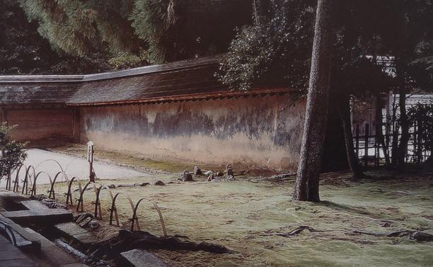A photograph of the "back garden" at Ryōan-ji Temple, Kyoto, the "Temple of the Dragon at Peace". 

Original image can be found on p. 23 of the late Christopher Alexander's 2002 remarkable book, The Phenomenon of Life -- Book One of his four part masterwork, The Nature of Order. 

The image centers on a thick, angular, reddish-brown plastered wall with a robust gabled roof-like top. The wall is weathered, and it surrounds on two sides a section of the temple's #Zen garden.

In the right lower foreground is a broad undulating area, covered in what appears to be a thick carpet of pale green #moss, with a single sizeable tree growing out of this mossy zone. None of the tree's branches are visible, only its single straight but slightly rightward-leaning trunk. The tree also casts an obvious slight shadow to the right, but the light source is unknown. Shadows elsewhere suggest a late hour of the day, perhaps even night time.

Outside the garden wall is bushy vegetation, and the branches of other trees, leaning over the wall in the darkness. In the lower left corner there are some blocks of stone or concrete, which may be the remnants of an old pathway, and a ribbony row of metal hoop-like forms.

This portion of the #Zen garden is not manicured or perfect in any usual sense. And yet, the atmosphere here is one of profound tranquility and almost overwhelming beauty. The scene is mysteriously other-worldly. It produces an extraordinary effect -- for this viewer, anyway.