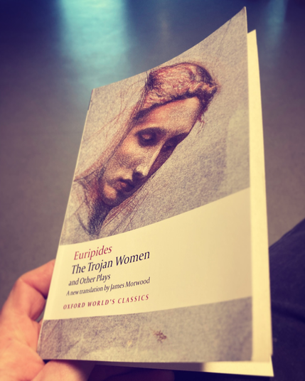 A photo of the front cover of Euripides’ Trojan Women. The shot is looking down into the lap of somebody holding the book and the studio floor can be seen in the background.