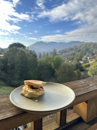 An English muffin breakfast sandwich with egg, bacon and cheese sits on a plate on a ledge, with Mt. Tamalpais in the background. A light haze envelops the mountain and blue sky peeks from behind the wispy clouds.