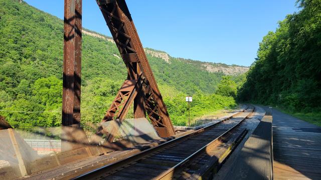 A paved bike trail follows a rail line across a rusty metal bridge as it cuts through a valley marked with high stone cliffs. The sky is a perfect shade of blue with no clouds visible.