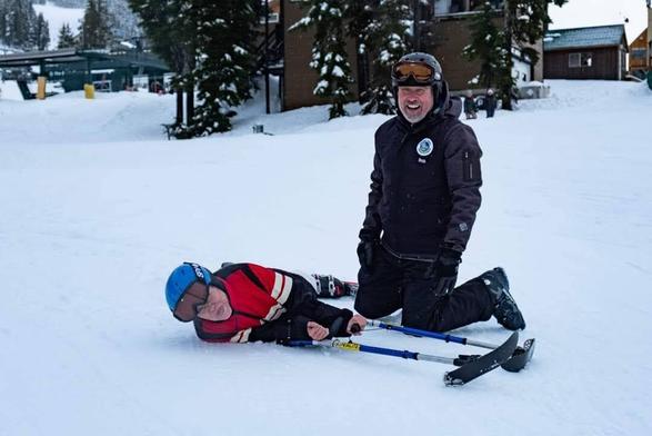 My adaptive ski instructor, kneeling beside me, in snow. I'm laying down sideways, holding 4 outrigger ski poles. We're both laughing.