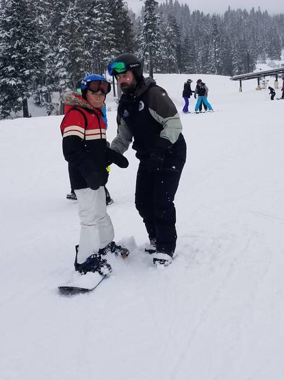 On a snowboard, my instructor is holding my gloved hand.  We are looking forward.  People on skis & snowboards are behind us.