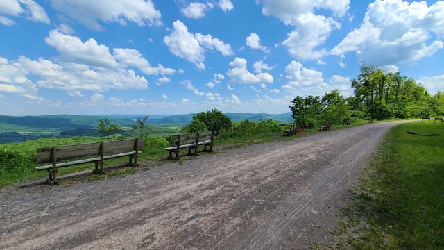A wide bike trail made of crushed gravel bends past a scenic overlook. The overlook has a view of a vast valley and a blue sky with a few scattered fluffy clouds.