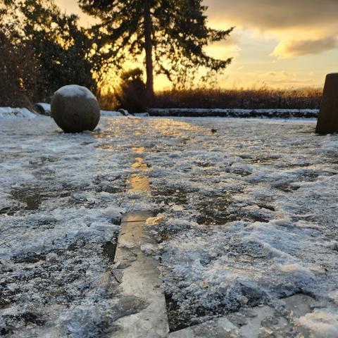 The sun, slightly obscured by a tree, in rough alignment with a silver colored metal strip, covered by ice and snow, pointing toward the setting sun.