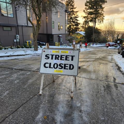 A sign that reads "Road Closed" stands at the top of an ice-covered hill in a residential neighborhood with townhouses and trees in the view.