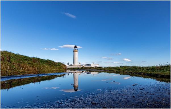 Lighthouse reflected in puddle on road. Clear blue sky.
