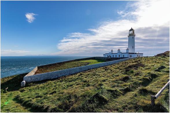 Imposing lighthouse . Wall surrounding garden on slope with see at back.