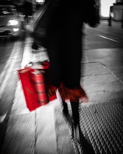 A mainly black and white image with only the reds highlighted. A woman is walking away from the camera carrying red xmas bags with the bottom of  their red dress is showing beneath their coat.