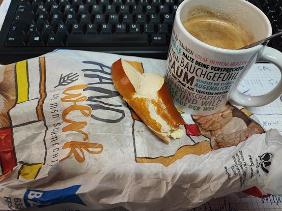 Butterbrezeln mit einem Pott Kaffee und einer Bäckertüte, in Hintergrund ist noch eine Tastatur zu sehen.