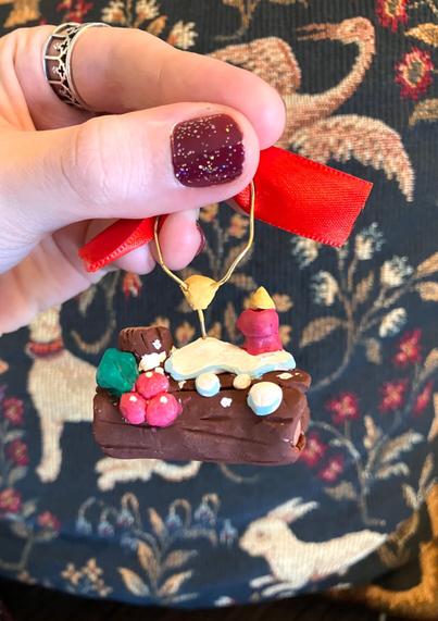 A woman’s hand holds up a yule log ornament hand-painted and made of sculpey clay and wire, in front of a French tapestry backdrop.