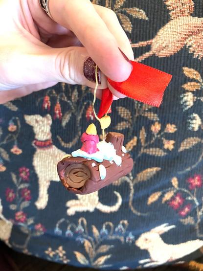 A woman’s hand holds up a yule log ornament hand-painted and made of sculpey clay and wire, in front of a French tapestry backdrop. A 3/4 view.