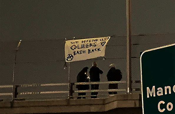 FAS comrades stand by on an overpass after hanging a banner that says "We defend us! Queers bash back!"