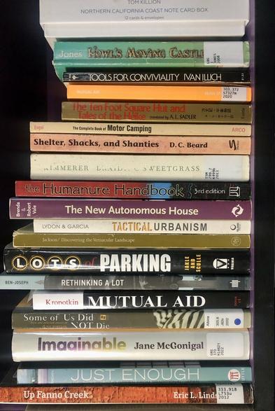 photo of bookshelf with 18 books: TOM KILLION NORTHERN CALIFORNIA COAST NOTE CARD BOX 12 cards & envelopes;  HOWL'S MOVING CASTLE; TOOLS FOR CONVIVIALITY IVAN ILLICH $1.25 MUTUAL AID DEAN S...;  The Ten Foot Square Hut Tales of the Heike translated by A. L. SADLER;  The Complete Book of Motor Camping; Shelter, Shacks, and Shanties, by D.C. Beard; BRAIDING S WEETGRASS; The Humanure Handbook, 3rd edition; The New Autonomous House; LYDON & GARCIA TACTICAL URBANISM; J.B. Jackson/ Discovering the Vernacular Landscape; Lots of Parking; BEN-JOSEPH, RETHINKING A LOT; Kropotkin MUTUAL AID Some of Us Did Not Die; Imagnable, bt Jane McGonigal; JUST ENOUGH; Up Fanno Creek