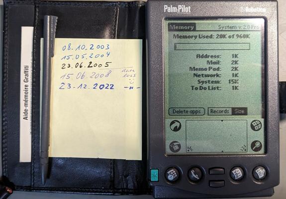 A PalmPilot. From left to right the top of a cheat sheet reads "Aide-mémoire Graffiti", the grey plastic touch-screen pencil, a post-it note with 5 dates indicating when the batteries were swapped last (08.10.2003, 15.05.2004, 22.06.2005, 15.06.2008 (data loss :-)), 23.12.2022, and on the far right the PalmPilot showing the Memory status and "System v.2.0. Pro" on the screen. On the top of the case PalmPilot is printed on the left and US Robotics on the right.