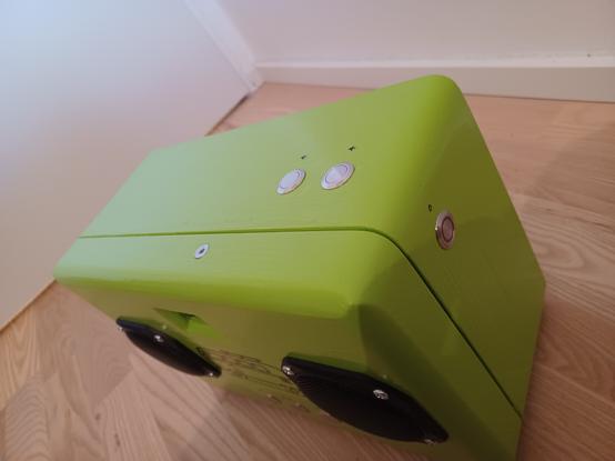 Phoniebox in grün in perspektivischer Aufnahme. Oben Laut-/Leiser-Button, seitlich Power-Button